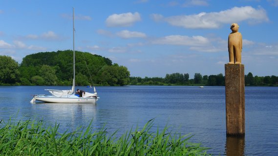 Blick auf den sommerlichen Vörder See mit einem ruhenden Seemann auf dem Segelboot und einem Kunstwerk mit dem Namen Seemann im Wasser