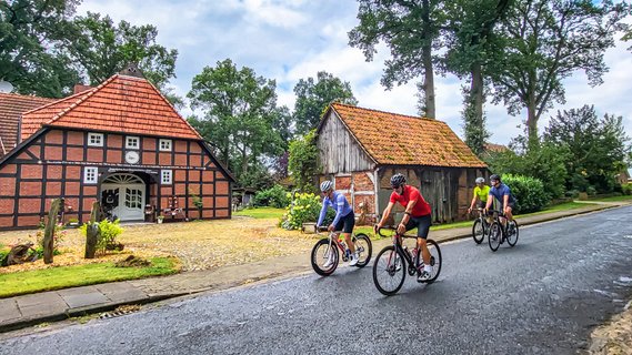 Vier Rennradfahrer fahren durch ein Dorf, im Hintergrund sind ein großes Fachwerkhaus und eine alte Fachwerkscheune zu sehen