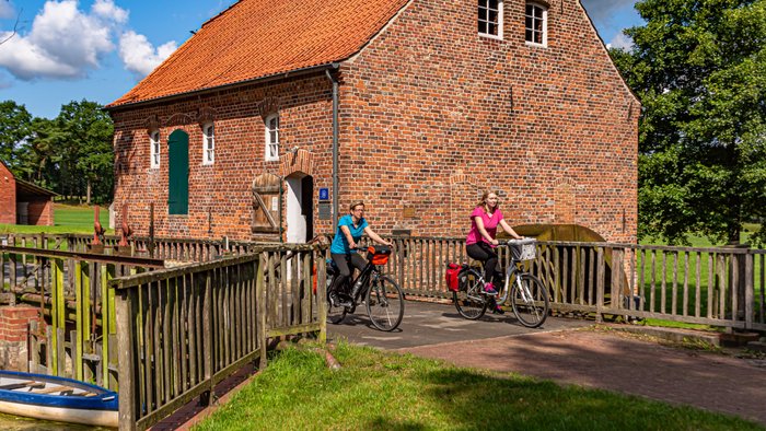 Zwei junge Radlerinnen fahren über die Brücke vor einer hitsorischen Wassermühle aus Backstein