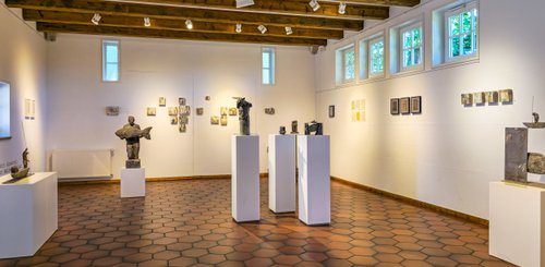 Im Raum stehen verschiedene Skulpturen auf weißen Podesten, während die Wände mit Kunstwerken geschmückt sind.