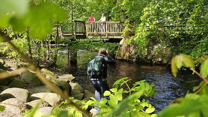Ein Fotograf steht an dem Bach Mehde inmitten von Bäumen und fotografiert ein Paar mit Fahrrädern auf der Brücke