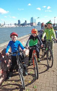 Eine Frau mit zwei Kindern schaut auf Fahrrädern sitzend in die Kamera, im Hintergrund ist die Elbphilharmonie in Hamburg zu sehen