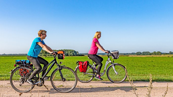 Zwei Radlerinnen unterwegs in einer weiten Landschaft mit grünen Feldern und blauem Himmel
