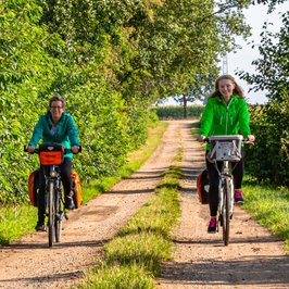 Zwei Radfahrerinnen sind auf dem Fahrrad unterwegs zwischen Wald und Feldern
