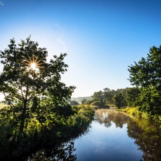 Die Landschaft mit grünen Bäumen, hinter denen die Sonne scheint, und blauem Himmel spiegel sich in der Wümme