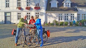 Eine dreiköpfige Familie hält auf einer Radtour vor einem historischen weißen Gebäude an, um sich auf einer Karte zu orientieren