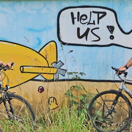 Zwei Radfahrer vor einem Graffiti mit einem gelben U-Boot und einer Sprechblase "Help us"