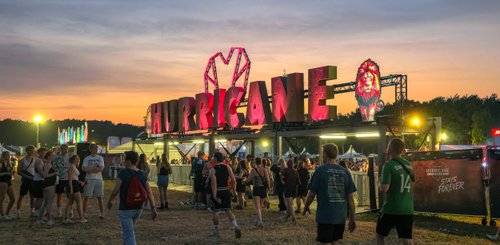Die bunten Lichter des Hurricane Festivals leuchten in der Dämmerung, während Menschen vor dem Eingang stehen und sich mit Freunden unterhalten.