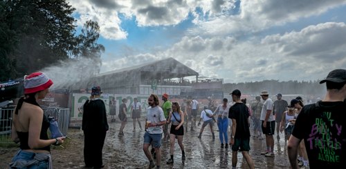 Eine belebte Festivalumgebung mit tanzenden Menschen im Schlamm, während ein Sprühnebel für Erfrischung sorgt.