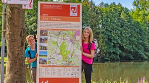 Zwei fröhliche junge Frauen schauen hinter einer Infotafel der NORDPADE hervor