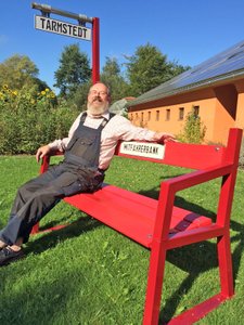 Herr Franke sitzt, mit einer Latzhose und grinsend, auf einer roten Mitfahrerbank in Buchholz