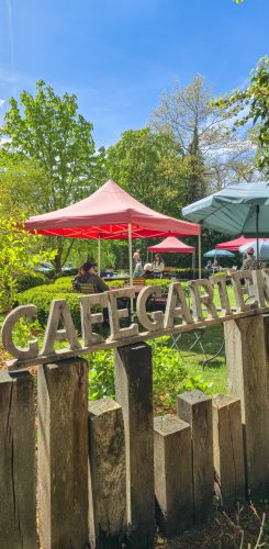 Die Holzschilder mit der Aufschrift 'Cafégarten' stehen im Vordergrund, während Gäste in der Sonne entspannen.