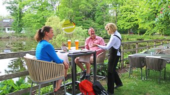 Ein Paar mit Wanderausstattung rastet an einem Teich, eine freundliche Bedienung lecker aussehende Speisen und Getränke gebracht