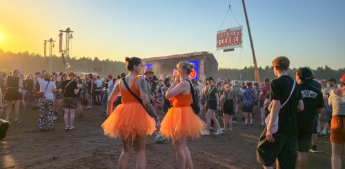 Zwei Frauen in orangefarbenen Tüllröcken stehen lächelnd in einer Menschenmenge bei einem Musikfestival im Sonnenuntergang.