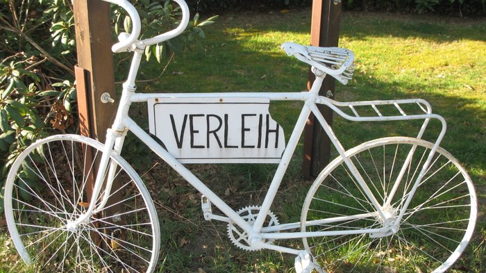 Ein Weißes Deko-Fahrrad mit einem Schild "Verleih" lehnt gegen einen Baum