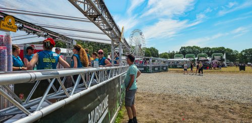 Eine Gruppe von Menschen, die an einem Getränkewagen stehen, während im Hintergrund Festivalbühnen und Zuschauer zu sehen sind.
