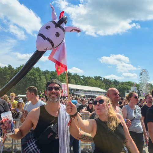 Eine fröhliche Person mit einer Giraffen-Puppe posiert vor einer Menge während eines Festivals unter strahlend blauem Himmel.