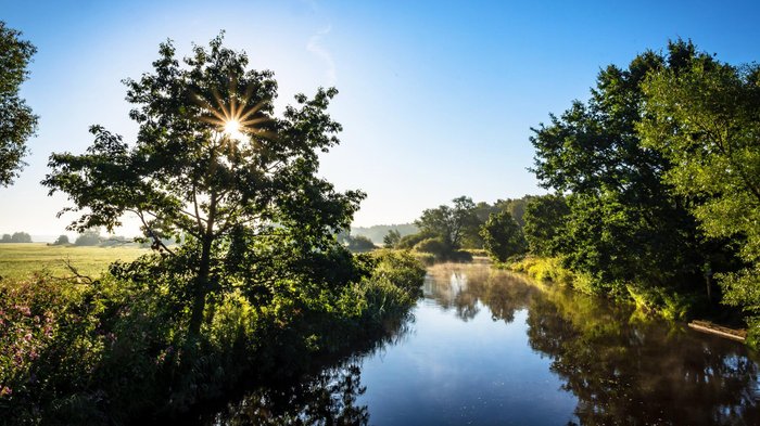 Die Landschaft mit grünen Bäumen, hinter denen die Sonne scheint, und blauem Himmel spiegel sich in der Wümme