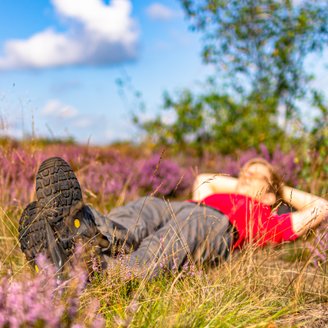 Eine Frau mit rotem T-Shirt und Wanderstiefeln liegt entspannt in einer lilafarbenen blühenden Heidefläche