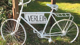 Ein Weißes Deko-Fahrrad mit einem Schild "Verleih" lehnt gegen einen Baum