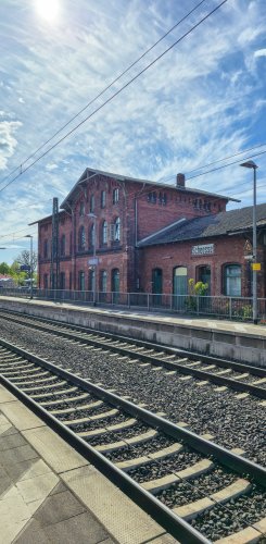Die Gleise am Bahnhof Scheeßel verlaufen in Richtung des eindrucksvollen, roten Bahnhofsgebäudes unter blauem Himmel.