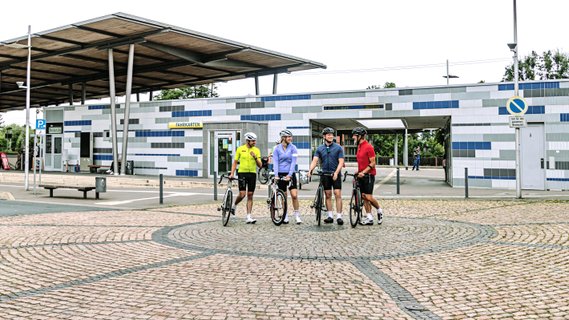 Vier Rennradfahrer stehen auf dem Kreisel vor dem modernen, in Blau und Grau gestalteten Bahnhof in Rotenburg (Wümme), bevor die Tour startet