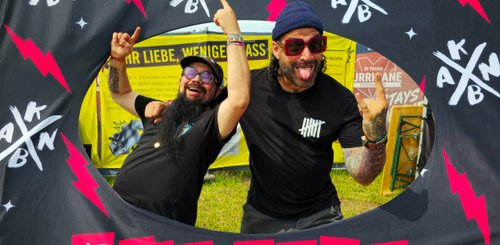 Mit Sonnenbrillen und witzigen Gesichtern stehen zwei Männer vor einem Festivalbanner und drücken Freude aus.