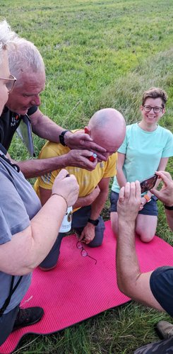 Udo und Petra hocken auf einer Matte, dabei wird Udo von Miro und seiner Frau einer Feuertaufe unterzogen