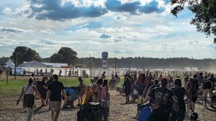 Festivalteilnehmer mit Rucksäcken und Wagen gehen über ein Feld, um Zelte und Bühnen im Hintergrund zu erreichen.