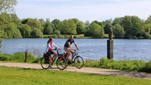 Ein Paar radelt im Frühling am Vörder See entlang, im See ist das Kunstwerk Seemann zu sehen