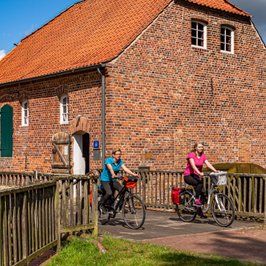 Zwei junge Radlerinnen fahren über die Brücke vor einer hitsorischen Wassermühle aus Backstein