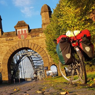 Ein Fahrrad mit rot-weißem Reisegepäck steht vor der Alten Harburger Elbbrücke, das von der Abendsonne in warmes Licht getaucht ist