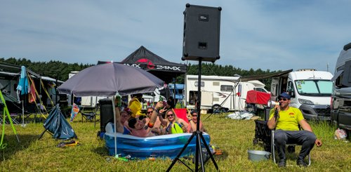 Eine Gruppe von Menschen sitzt in einem Planschbecken, genießt Getränke und hat Spaß auf einem Festivalgelände.
