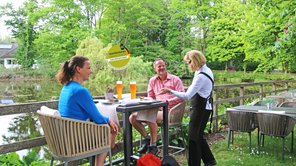 Ein Paar mit Wanderausstattung rastet an einem Teich, eine freundliche Bedienung lecker aussehende Speisen und Getränke gebracht