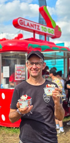 Ein Mann hält zwei Eisbecher mit Erdbeeren und Sahne, vor einem bunten Verkaufsstand mit 'GLANTZ TO GO'.