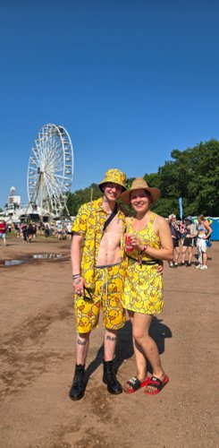 Ein freudiges Paar lächelt in bunten Entenkostümen vor einem Riesenrad auf einem Festivalplatz.