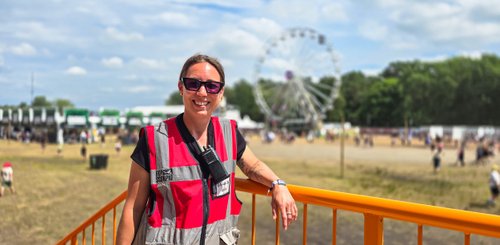 Eine lächelnde Person in einem roten Sicherheitsanzug steht auf einer Plattform, während im Hintergrund das Riesenrad dreht.