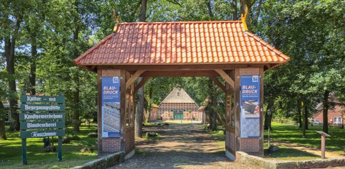 Ein einladendes Tor mit rotem Ziegeldach führt in den gepflegten Garten des Heimatmuseums unter grünen Bäumen.