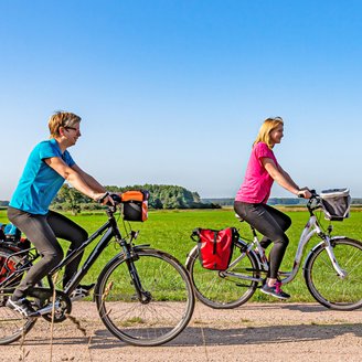 Zwei Radlerinnen unterwegs in einer weiten Landschaft mit grünen Feldern und blauem Himmel