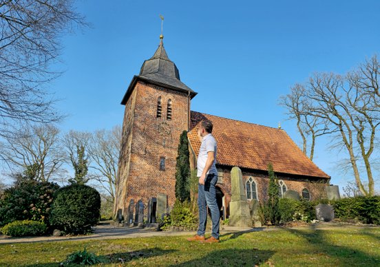 Ralph zeigt auf die Turmuhr der Oeser Kirche in der Frühlingssonne