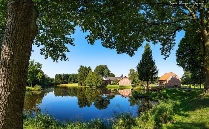 Der blaue Himmel spiegelt sich im Teich vor der Wassermühle Bademühlen sowie angrenzenden Backsteinhäusern