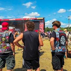 Festivalbesucher mit Metal-Kutten schauen auf die Bühne, während andere Besucher in der Menge sich unterhalten und entspannen.