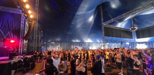 Teilnehmer eines Festivals, die in einem großen, blauen Zelt versammelt sind, genießen die Musik und die Atmosphäre.