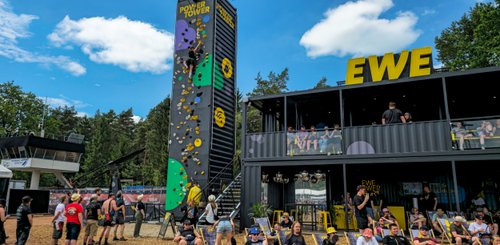 Ein Kletterturm mit bunten Griffen zieht die Aufmerksamkeit der Zuschauer an, während einige entspannte Besucher im Vordergrund sitzen.