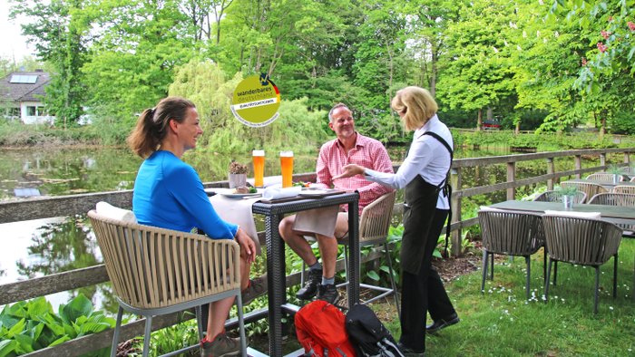 Ein Paar mit Wanderausstattung rastet an einem Teich, eine freundliche Bedienung lecker aussehende Speisen und Getränke gebracht