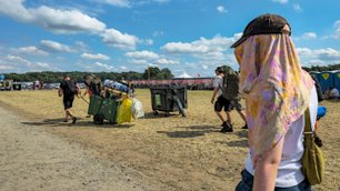 Drei Personen bewegen sich mit schwerem Gepäck über das trockene, staubige Festivalgelände unter einem klaren Himmel.