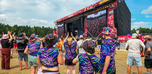 Gruppe von Festivalbesuchern, die in bunten Outfits und Hüten vor der Bühne jubeln und den Auftritt genießen.