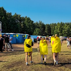Drei Menschen in bunten Bananenkostümen gehen fröhlich auf einem Festivalgelände mit blauen Toiletten im Hintergrund.