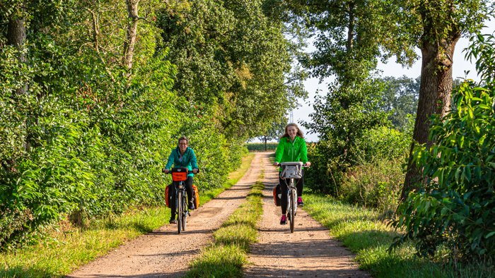 Zwei Radfahrerinnen sind auf dem Fahrrad unterwegs zwischen Wald und Feldern