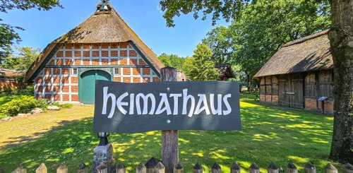 Die idyllische Szenerie zeigt das Heimathaus, eingerahmt von hohen Bäumen und klarblauem Himmel, ideal für Besucher.